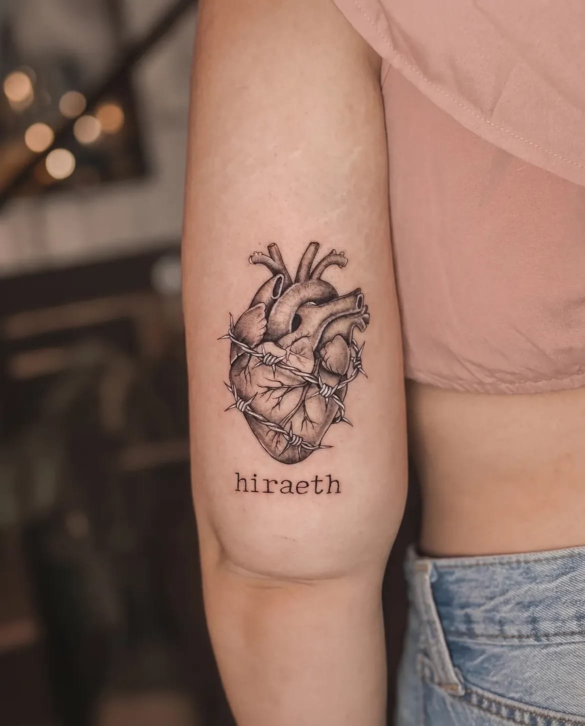Fine-line anatomical heart tattooed on a forearm above the lettering “hiraeth”.