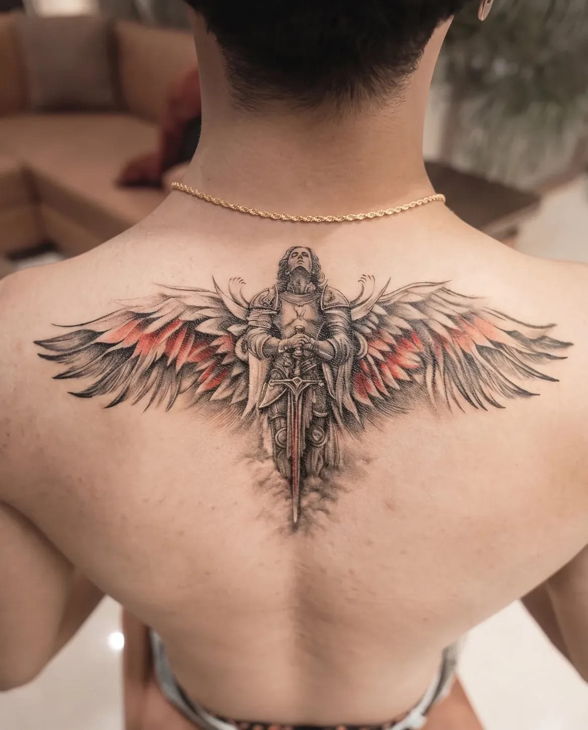 Large winged figure tattooed across an upper back in black, grey and red ink.